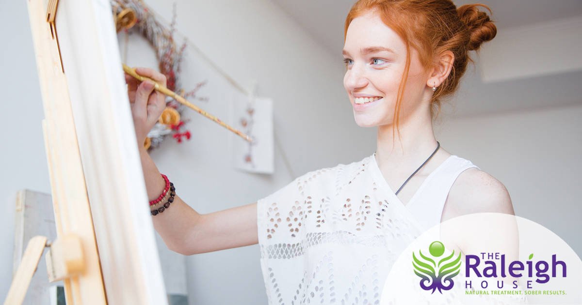 An attractive woman in a white lace top smiles as she paints a canvas.