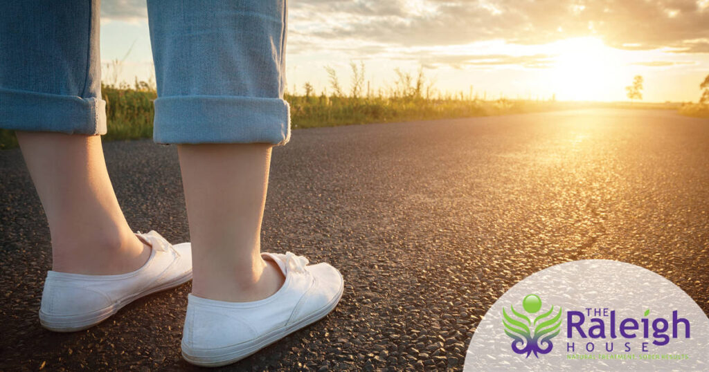 A woman stands on an asphalt road, with a long journey ahead of her as the sun shines on the horizon.