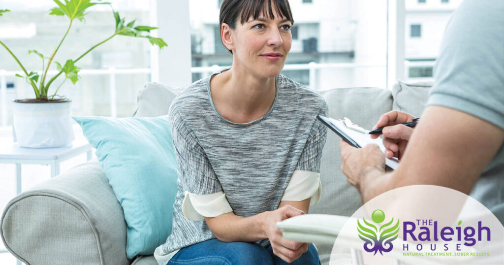 A woman sits on a couch in a sunny room, talking to her therapist about alcohol addiction as he takes notes.