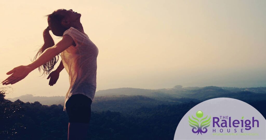 A young woman stands at the top of a mountain, her face tilted toward the sky and her arms thrown back in a gesture of pure joy.