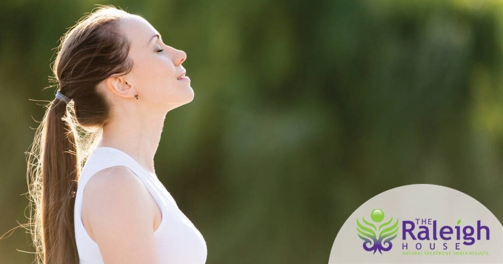 A young woman stands in a park, her face tilted toward the sun and eyes closed.