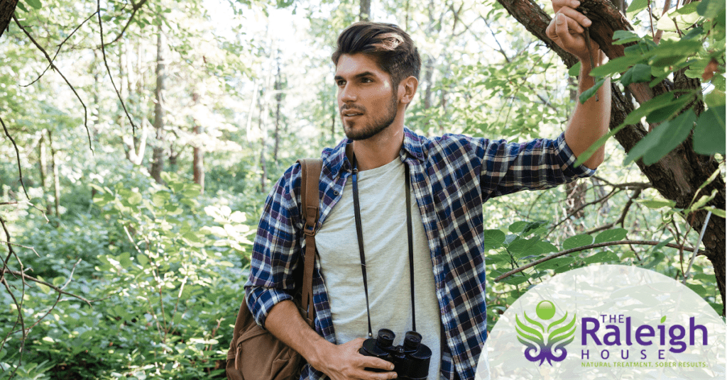 A young man with binoculars has a smile on his face as he spots something in a forest.