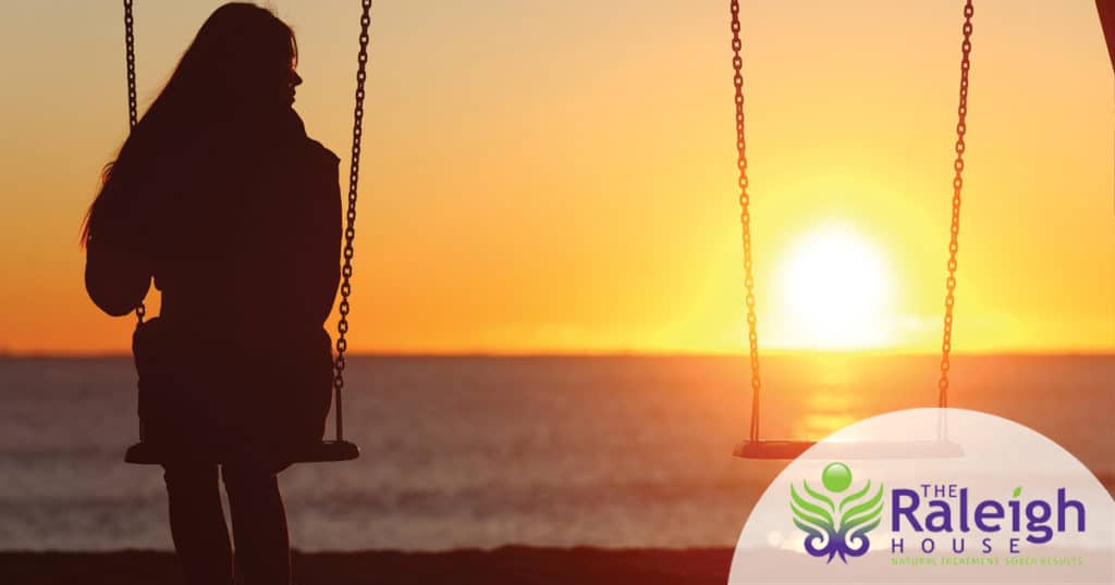 A young woman sits along on a swing at the beach as the sun sets behind her.