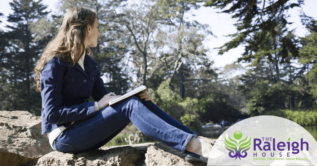 A young woman sits on a rock overlooking a lake and writes in her journal.