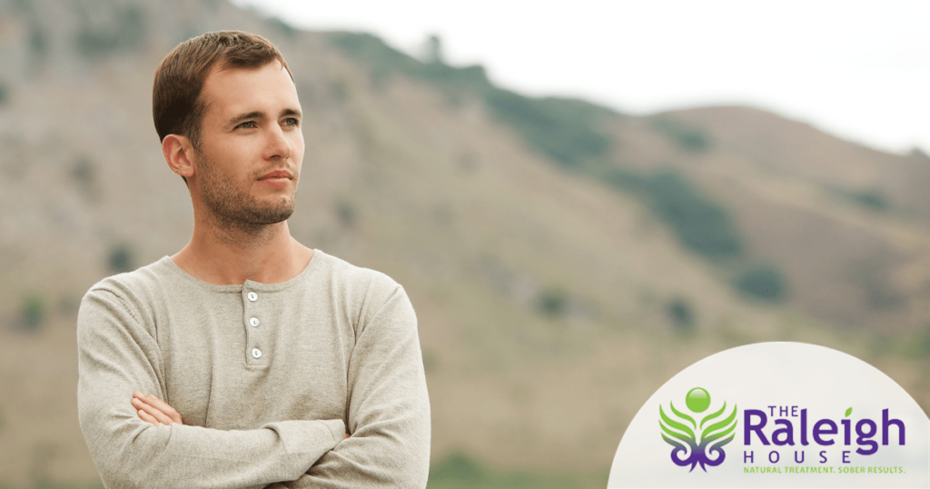 A clean-cut man with sandy brown hair stands with his arms crossed in front of a mountain. 