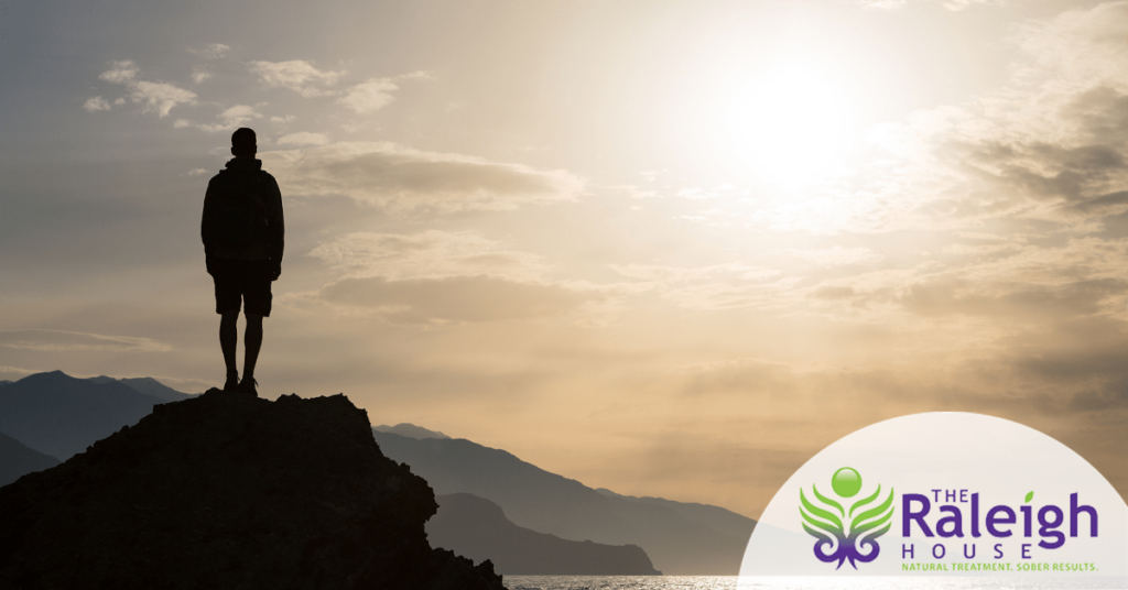 A man stands on a large rock overlooking a lake as the sun sets.