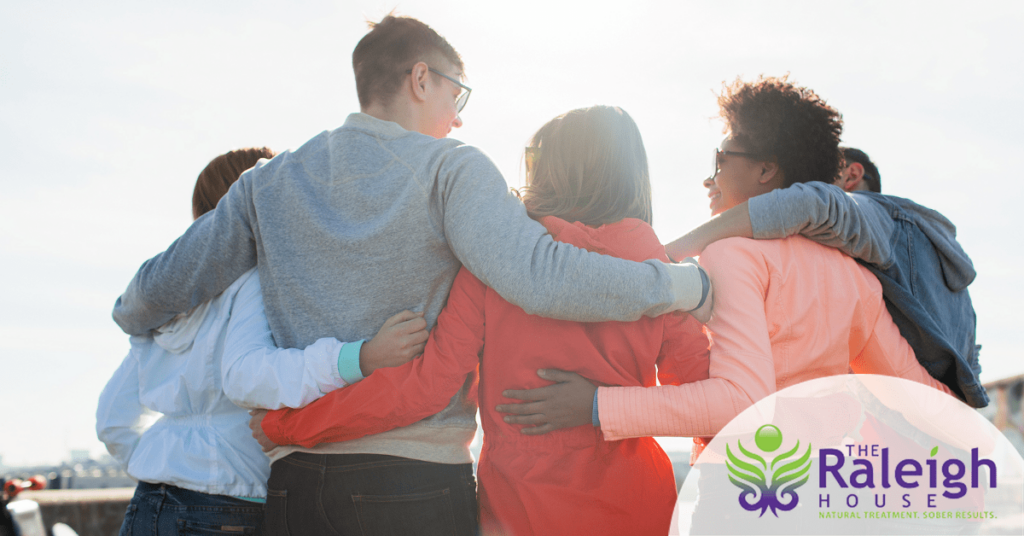 A group of five adults stands outside in a semi-circle with their arms around each other.