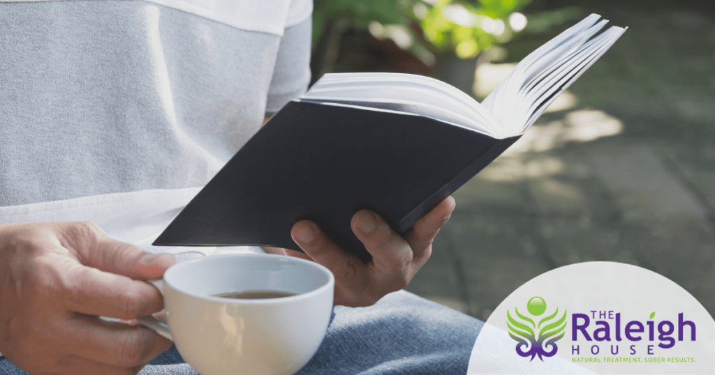 A close-up of a book being read by a man sitting outside by a tree with a cup of coffee.