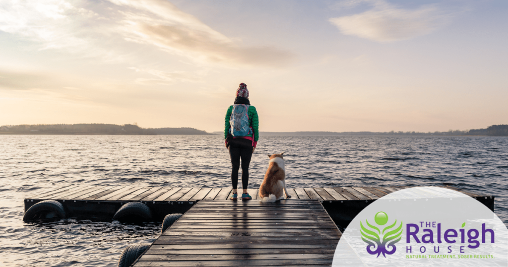 A woman stands at the end of a pier in the early morning light with her dog at her side.