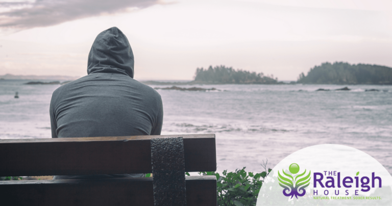 A man sits alone on a bench, looking off into the distance at a calm lake.