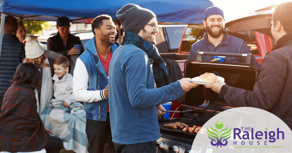 A group of people gather around a grill in a parking lot to tailgate before a football game. 