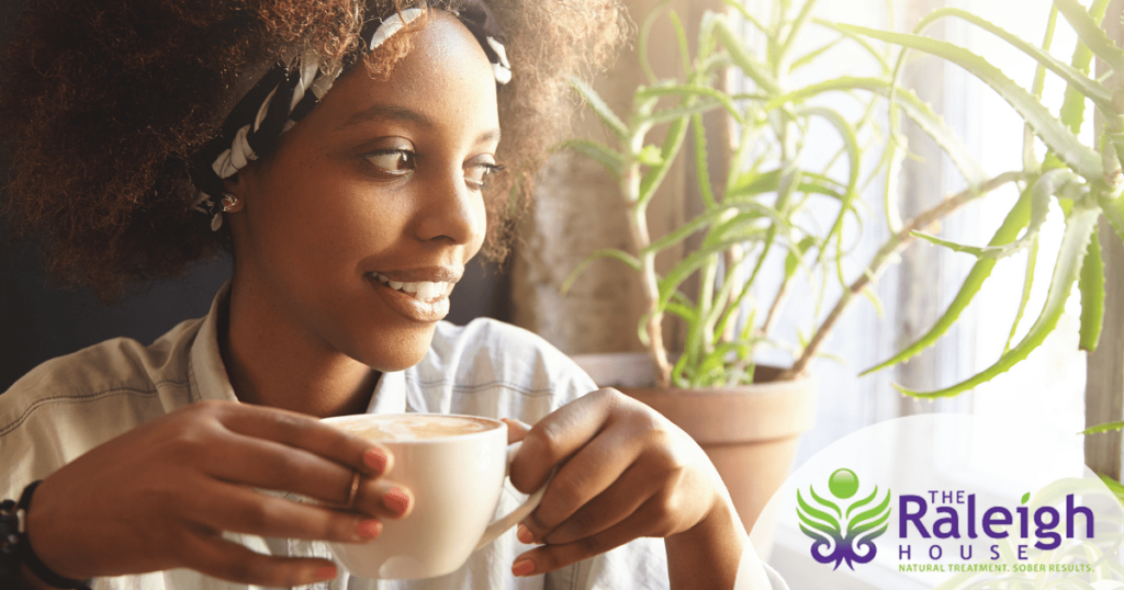 A woman smiles and drinks coffee as she looks out her apartment window.