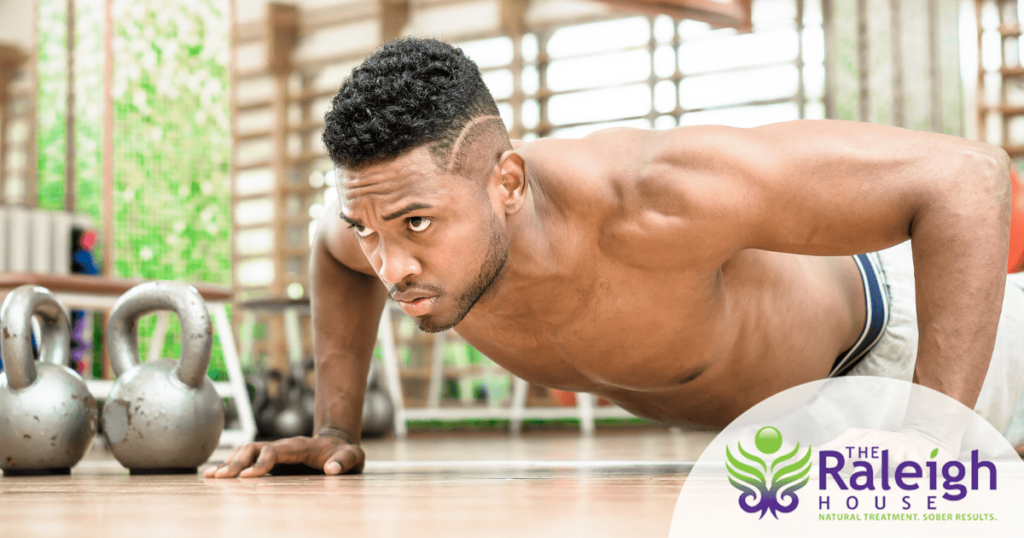 A young man does a plank in a gym, with kettle balls next to him. 