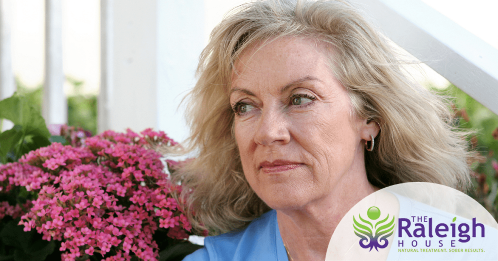 An older woman sits by some beautiful flowers.