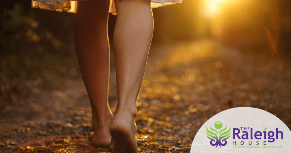 A barefoot woman wearing a cotton dress walks on a leaf-strewn dirt path toward the sunset. 