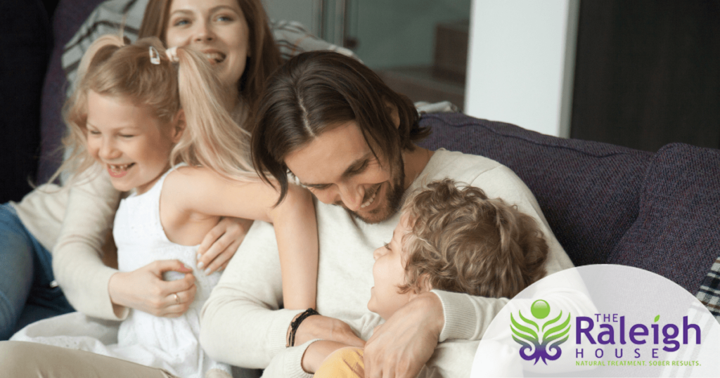 A mom, dad and their two young children laugh together on the couch.