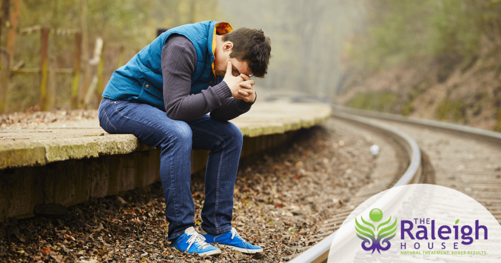 A sad-looking man sits by railroad tracks with his head in his hands.