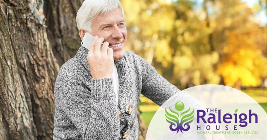 A mature man sits against the trunk of a tree while talking on the phone with a smile on his face.