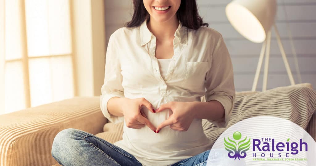 A pregnant young woman holds her hands in the shape of a heart on her belly while sitting cross-legged on a couch.