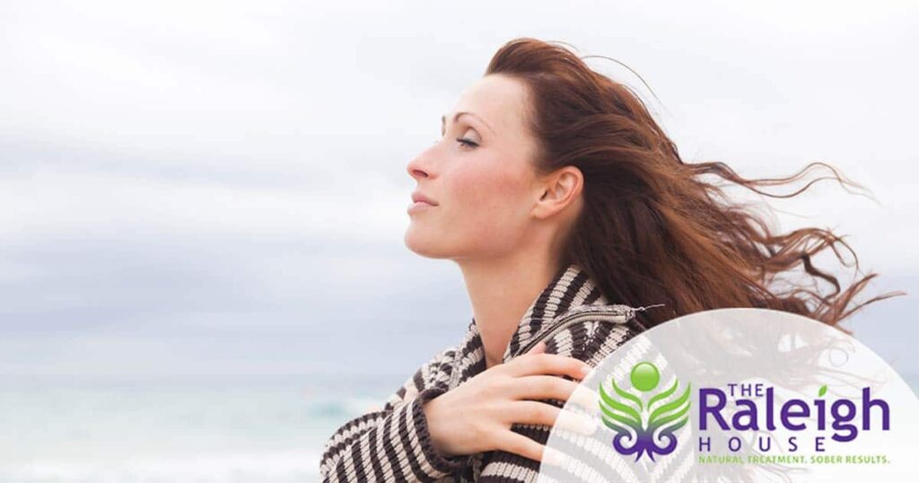 A woman wearing a sweater stands on the beach, looking into the distance as the wind blows her hair.