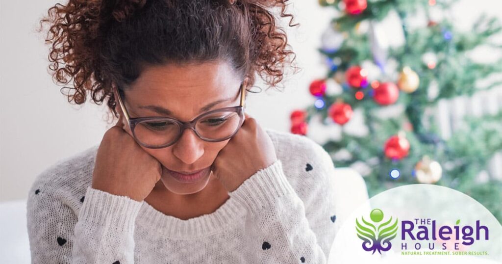 A young woman sits near a Christmas tree with a forlorn look on her face.