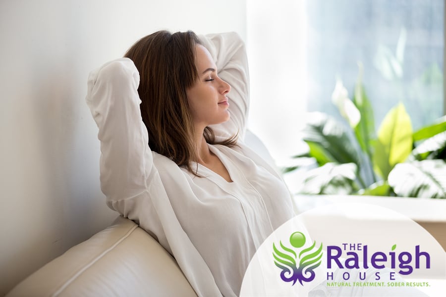 A woman sits on a sofa with her hands behind her head, looking relaxed and happy.