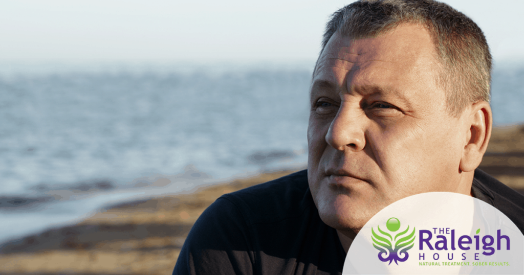 A middle-aged man has a serious look on his face as he stands with the ocean behind him.