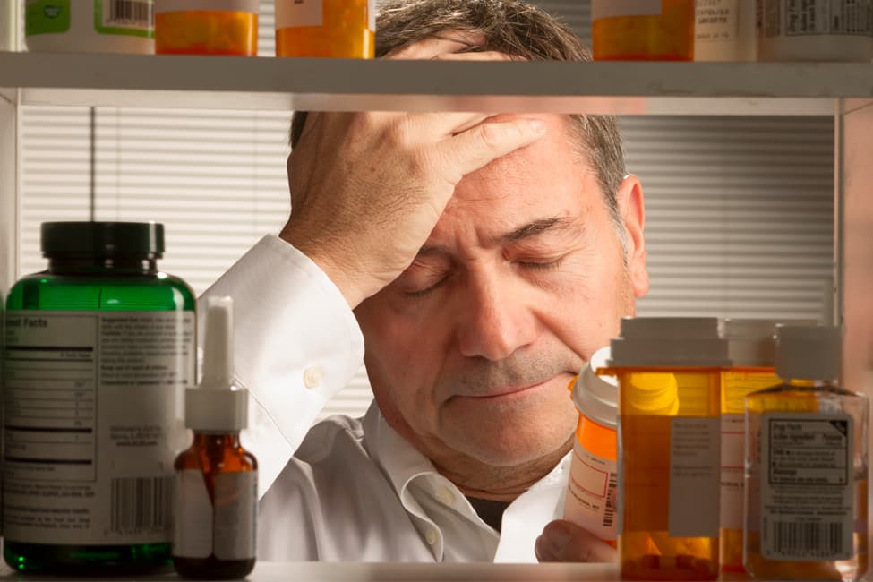 Man at medicine cabinet holding his head while reading a prescription pill bottle.