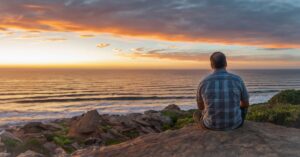 man sits by the ocean and watches the sunset.