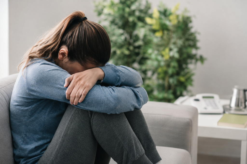 Woman sitting alone on a couch with her head down, showing signs of emotional exhaustion, depression, anxiety, and mental health burnout.